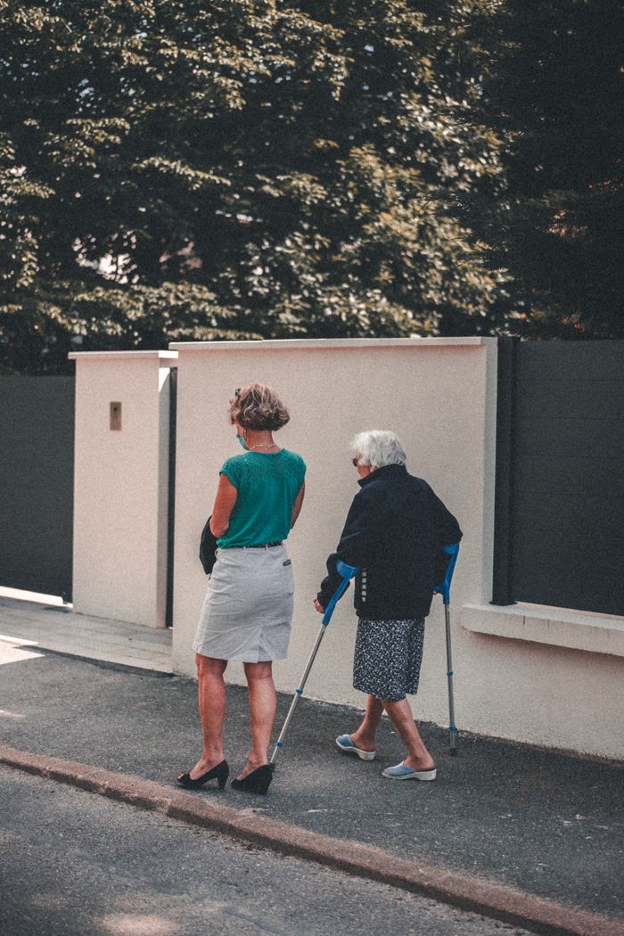 hero-contact Senior woman with crutches walking alongside a companion on a sunny day in Lyon, France.
