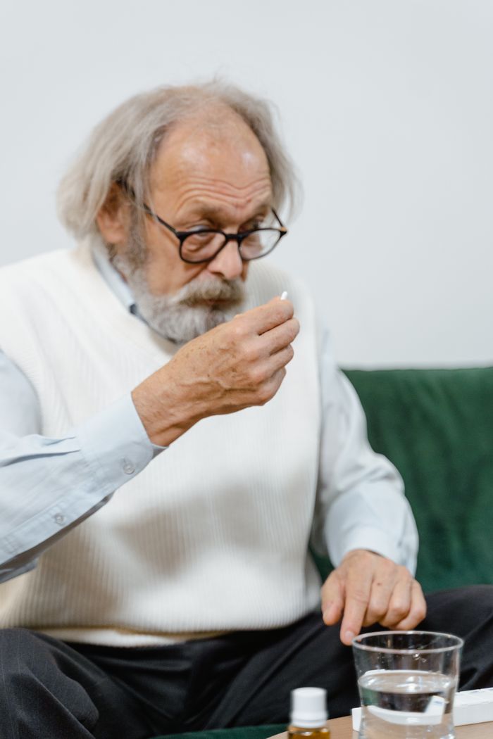 Elderly man with eyeglasses holding a pill, sitting on a couch indoors, ready to take his medication.